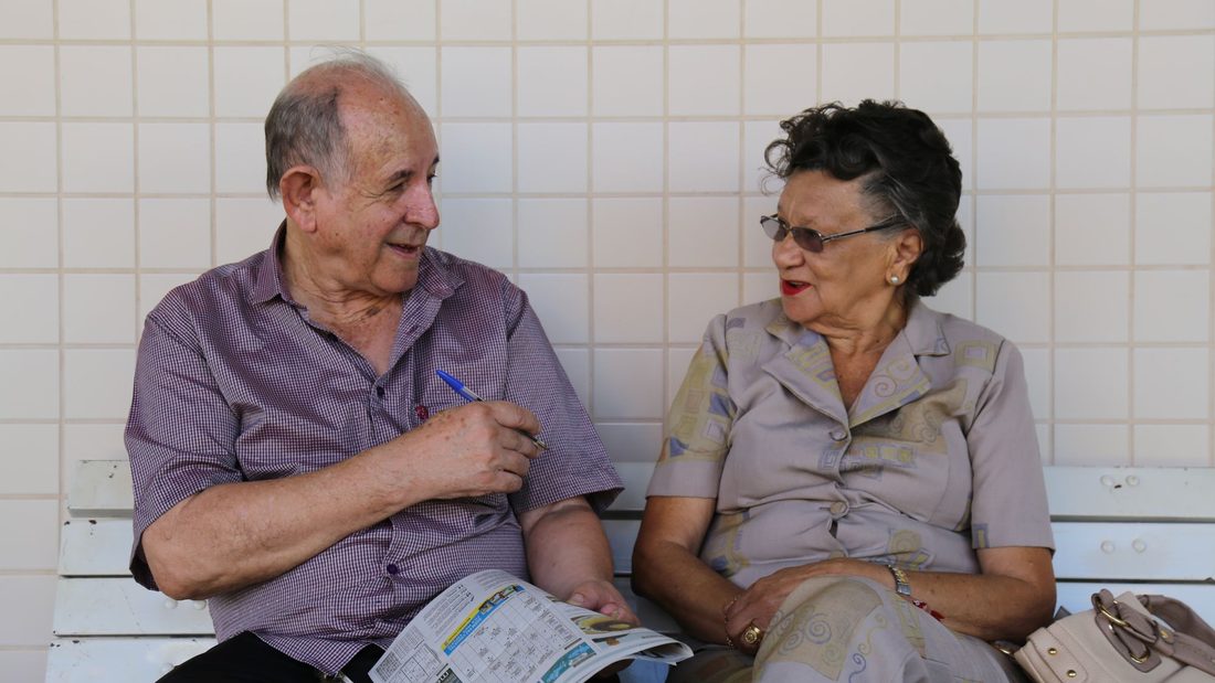 Casal de idosos conversando e sorrindo num banco de praça